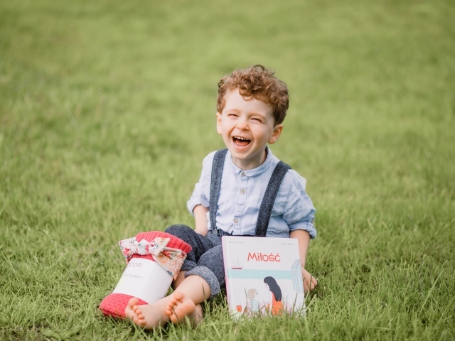 Маленький улыбающийся мальчик сидит на зеленой траве с книгой 