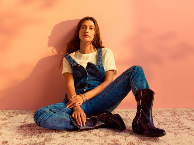 Американская модель Хейли Болдуин в джинсовом комбинезоне сидит у стены