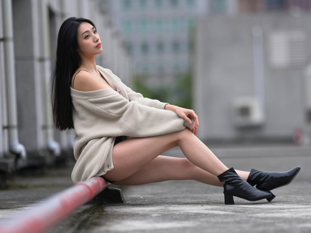 Грустная азиатка в теплой кофте сидит на трубе