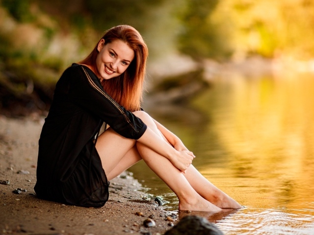 Улыбающаяся девушка в черном платье сидит у воды