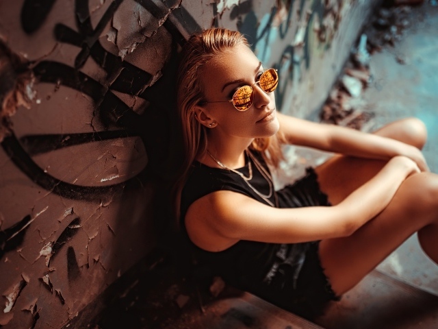 Молодая девушка в очках сидит у стены