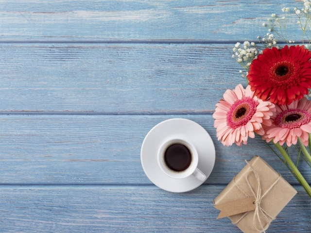 Три герберы с чашкой кофе и подарком на синем столе