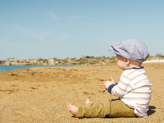 Маленький мальчик в кепке сидит на песке у воды