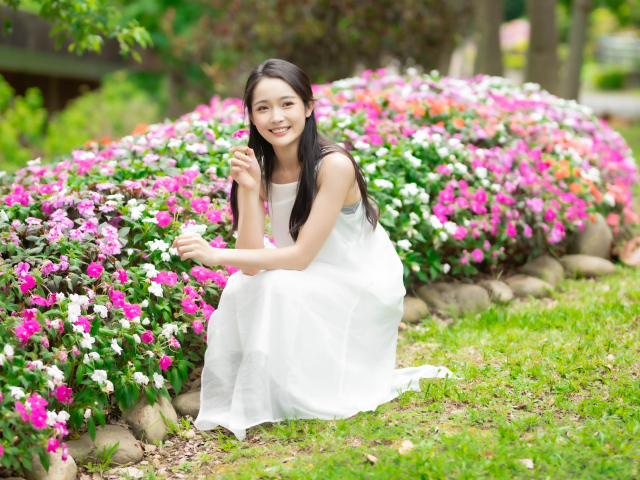 Милая улыбающаяся азиатка в белом платье сидит у цветов