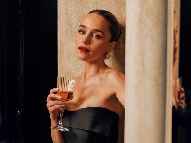 Актриса Эмилия Кларк с бокалом стоит у стены