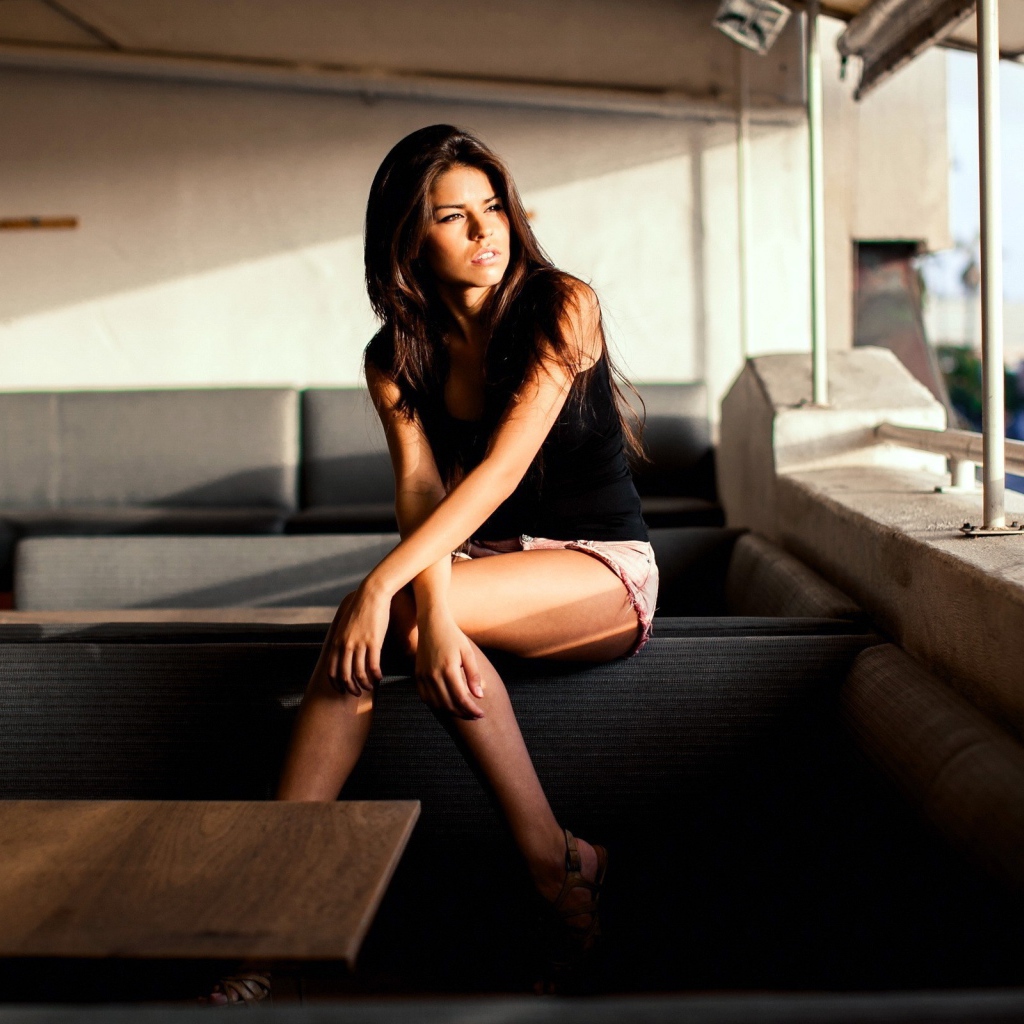 Брюнетка сидит на спинке дивана и смотрит в окно