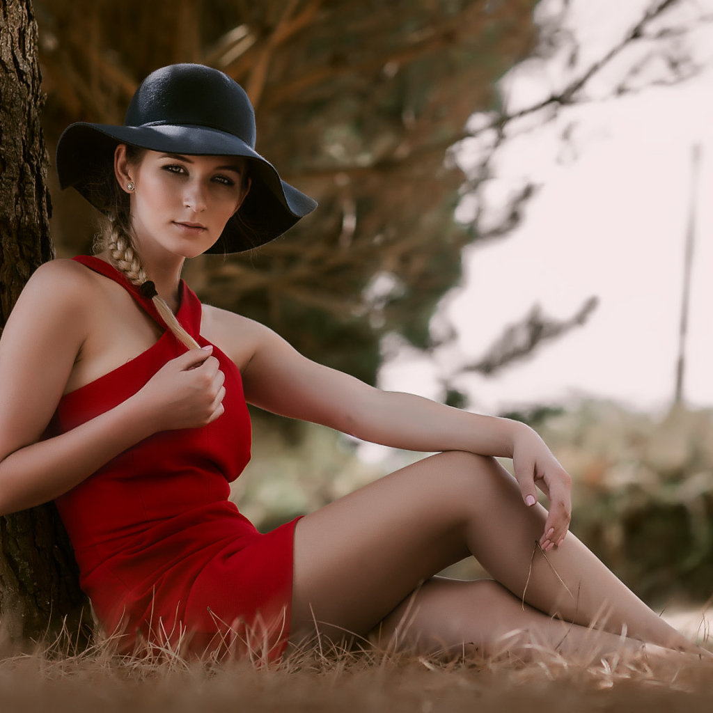 Красивая девушка в красном платье и черной шляпе сидит под деревом