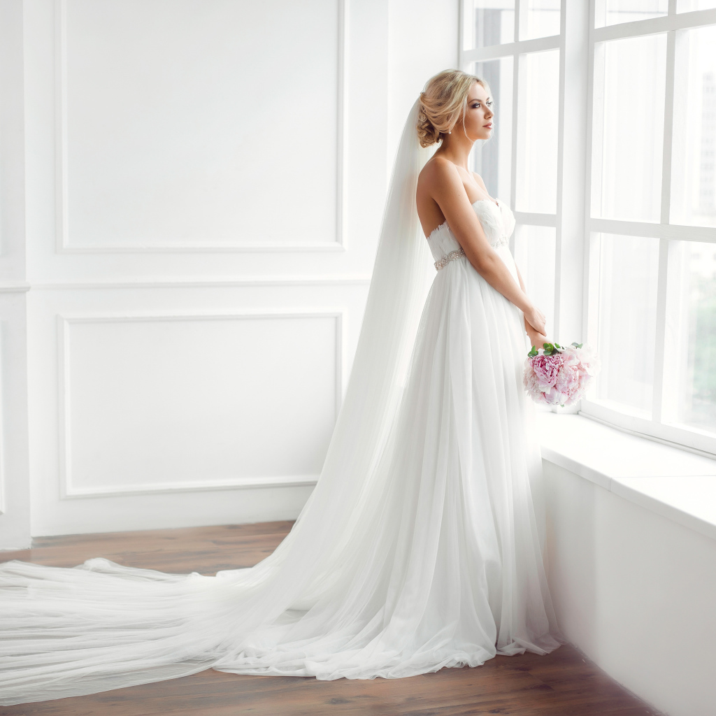 Красивая девушка в белом свадебном платье с букетом стоит у окна