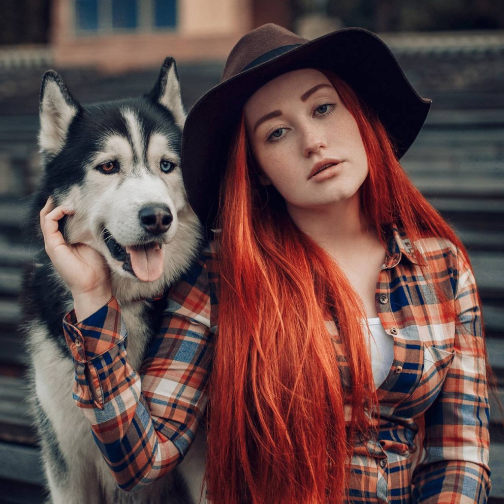 Рыжеволосая девушка в черной шляпе сидит с хаски