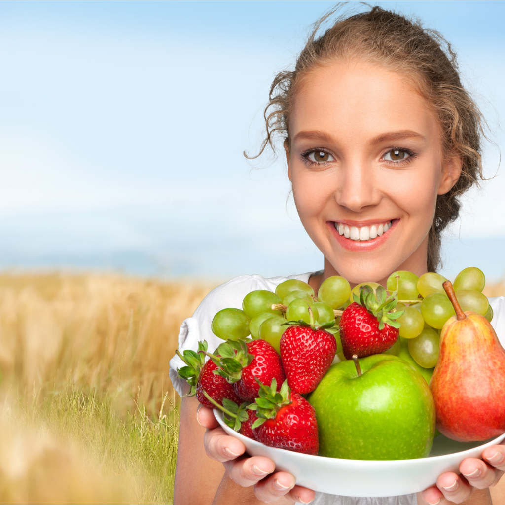 Улыбающаяся девушка шатенка с тарелкой с яблоками, виноградом, клубникой и грушей в руках