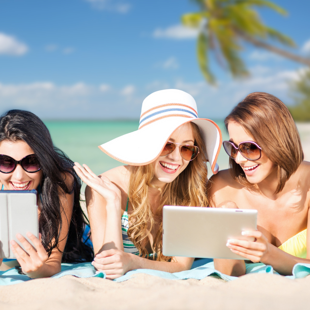 Три улыбающиеся девушки загорают на пляже с планшетами в руках