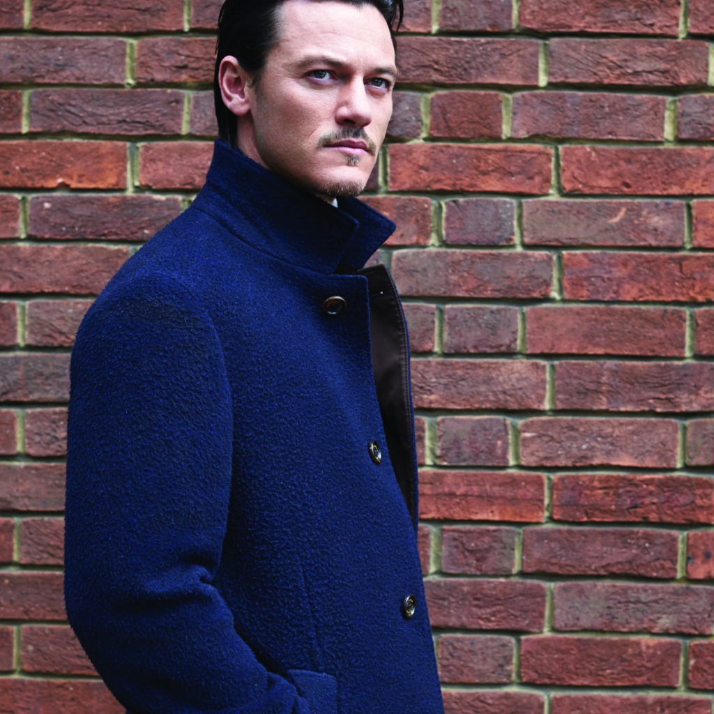 Актер Люк Эванс в синем плаще у кирпичной стены