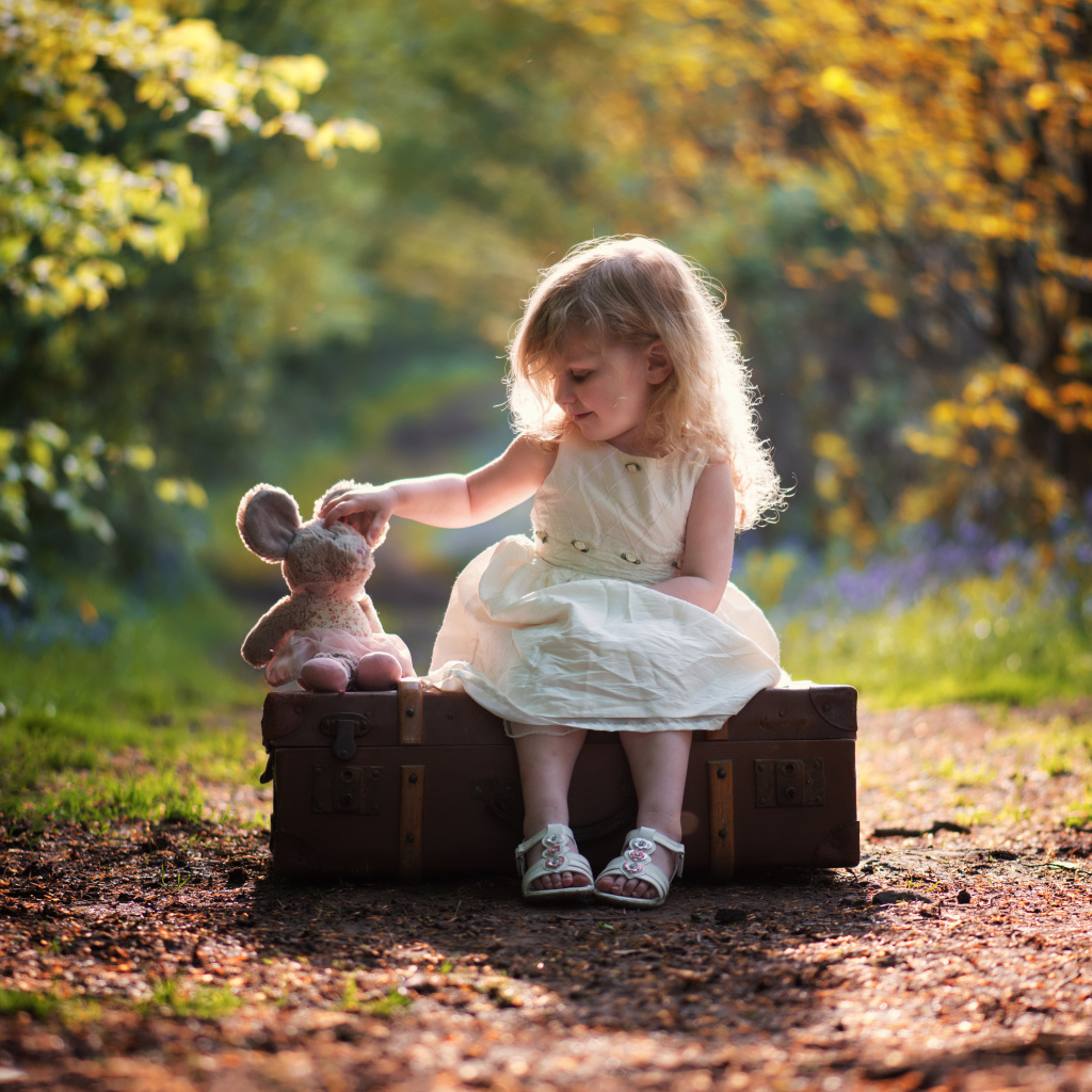 Маленькая девочка в белом платье сидит на большом чемодане с игрушкой