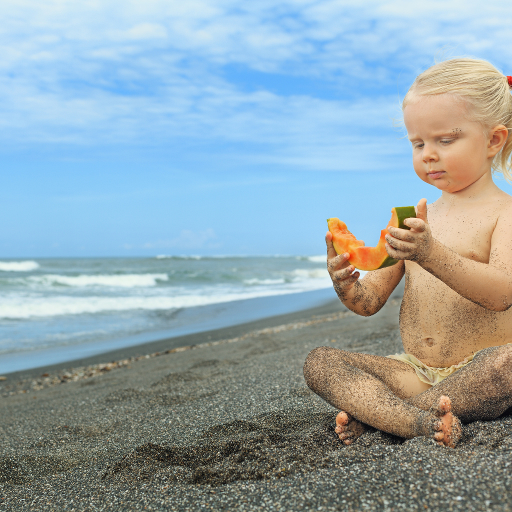 Маленькая девочка сидит на песке у моря с кусочком дыни