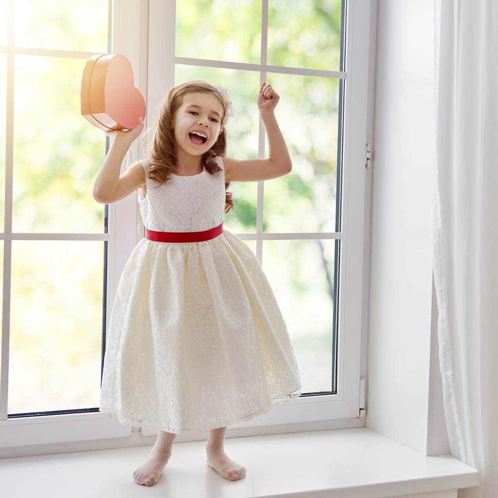 Маленькая девочка с коробкой в форме сердца в руках стоит на подоконнике