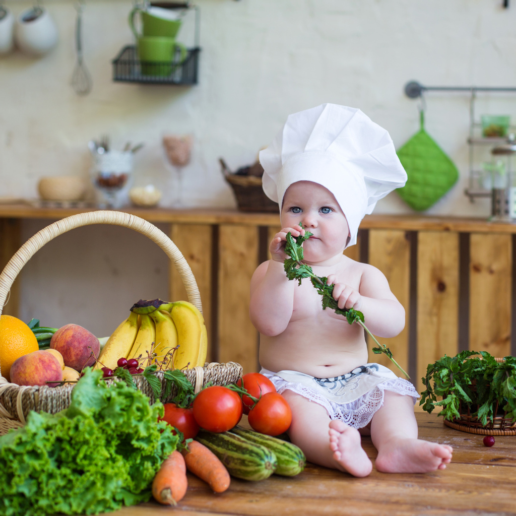Маленький ребенок в шапке повара сидит на столе с овощами