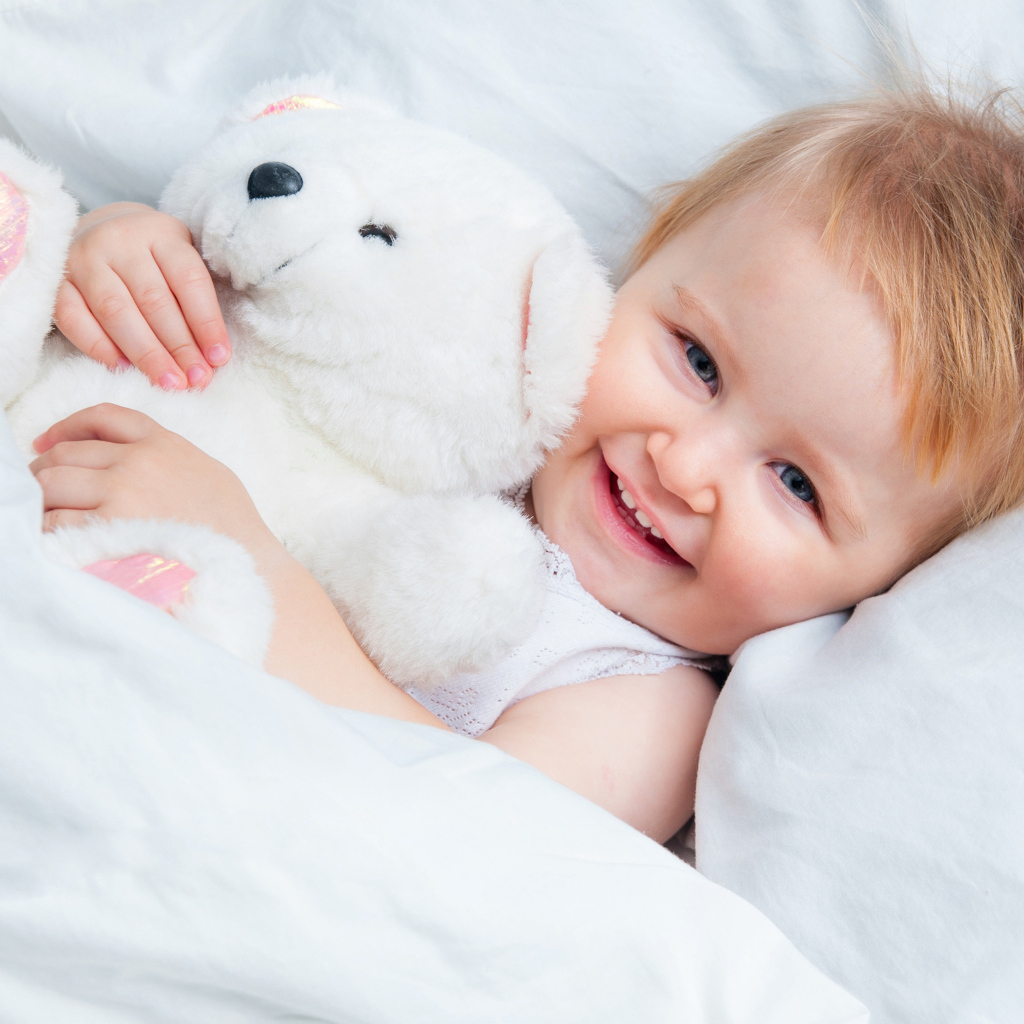 Улыбающийся ребенок лежит в кровати с белым плюшевым медвежонком