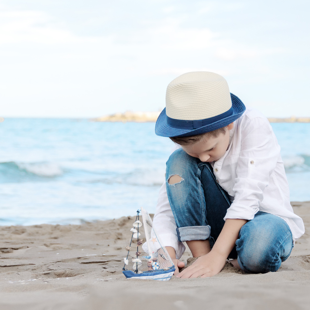 Маленький мальчик в шляпе играет с игрушечным корабликом на песке