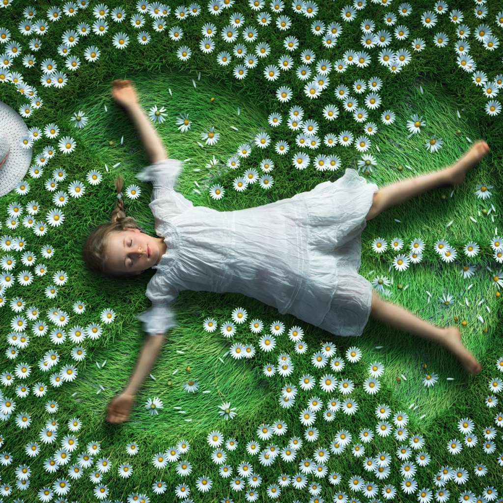 Маленькая девочка в белом платье лежит на зеленой траве с ромашками