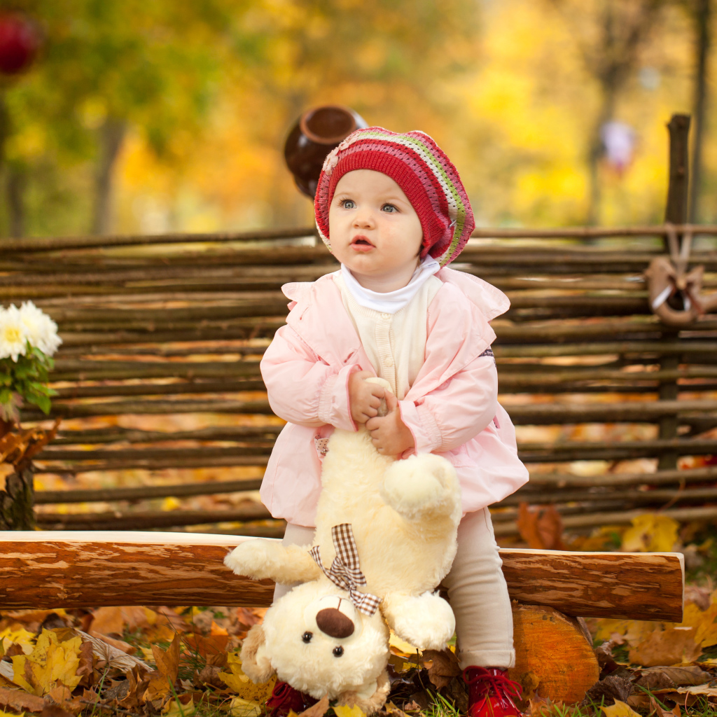 Маленькая девочка с плюшевым мишкой сидит на деревянной лавке осенью