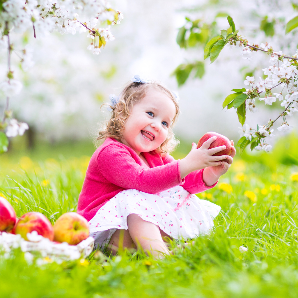 Маленькая улыбающаяся девочка сидит на зеленой траве с яблоками