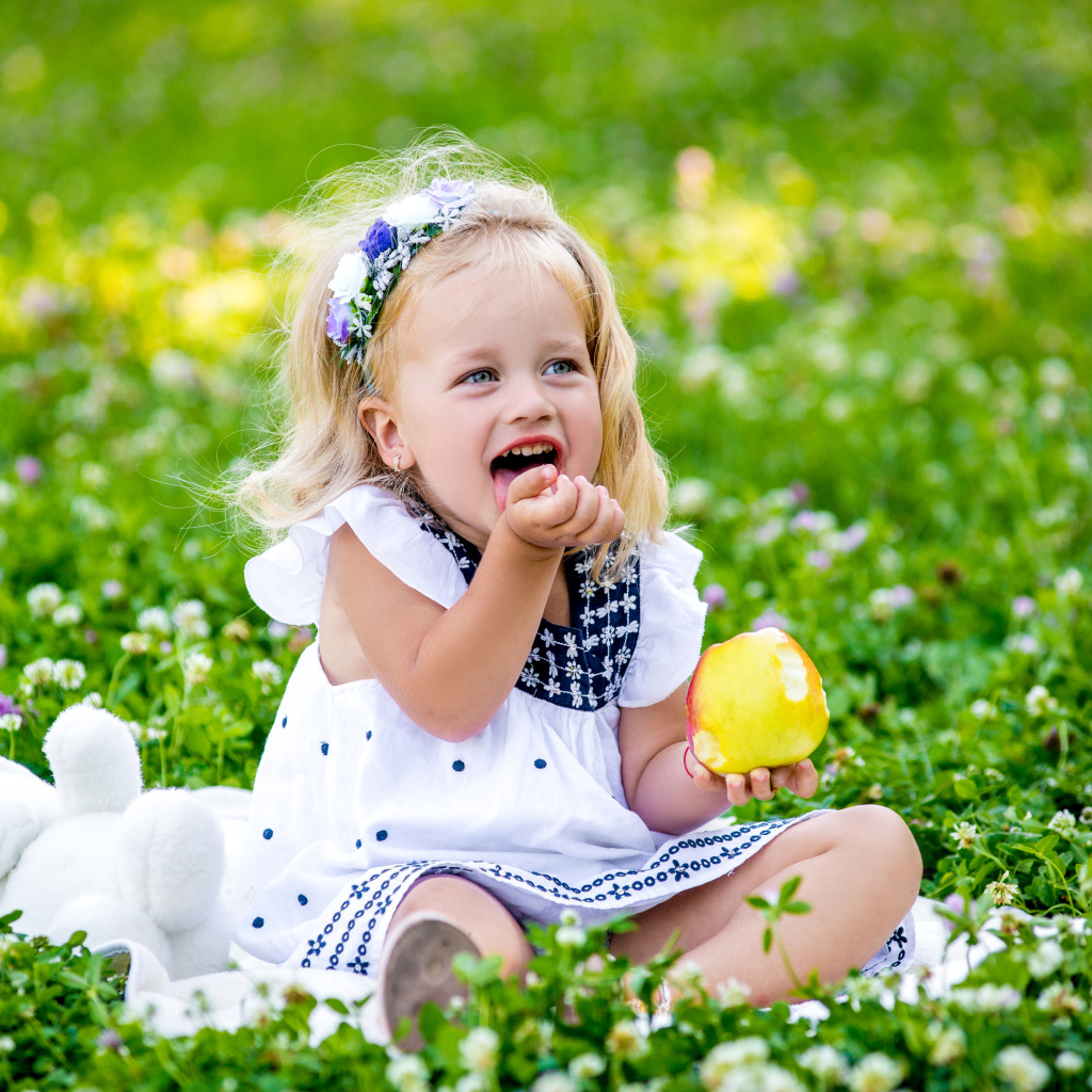 Маленькая улыбающаяся девочка с яблоком сидит на зеленой траве