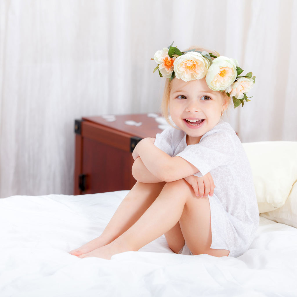 Улыбающаяся девочка с венком на голове сидит на кровати