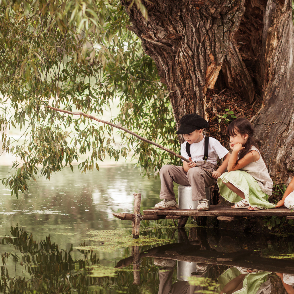 Две маленькие девочки и мальчик сидят на берегу пруда с удочкой
