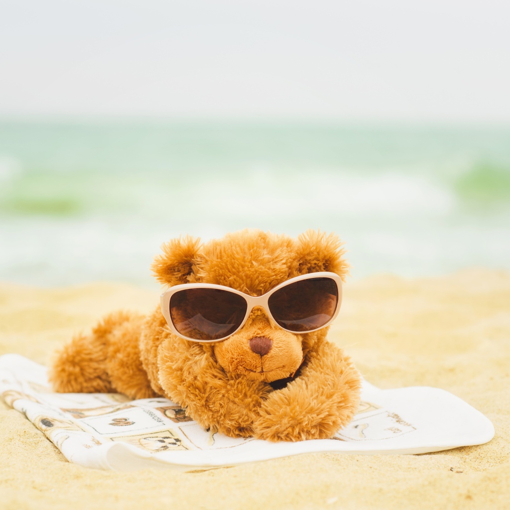 Плюшевый мишка в солнечных очках лежит на пляже
