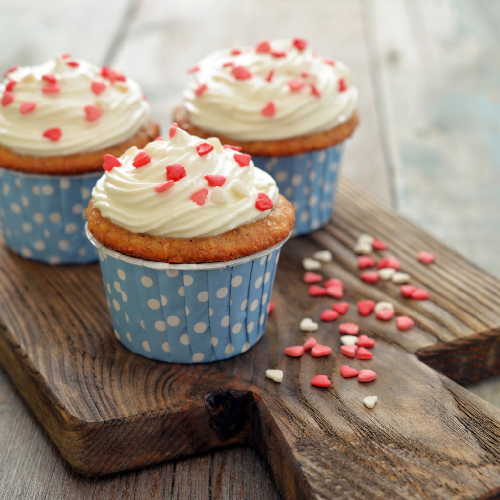 Три кекса со сливками украшены сердечками стоят на разделочной доске