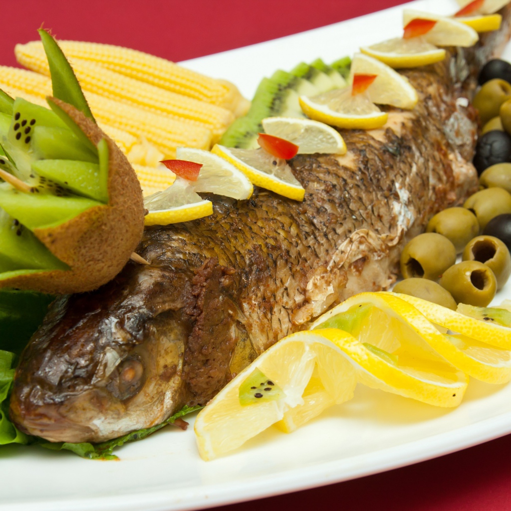 Запеченная рыба с оливками, лимоном, киви, кукурузой и листьями салата на белой тарелке