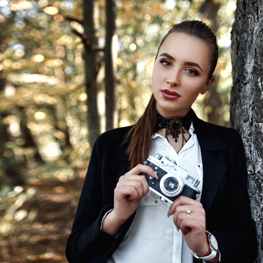 Красивая девушка с фотоаппаратом в руках стоит у дерева