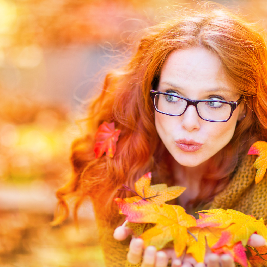 Рыжеволосая девушка в очках держит в руках опавшие листья 