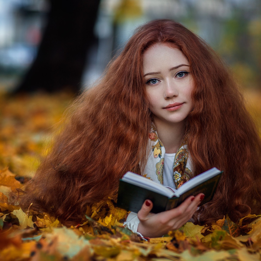 Рыжеволосая девушка с книгой лежит на опавших листьях осенью 