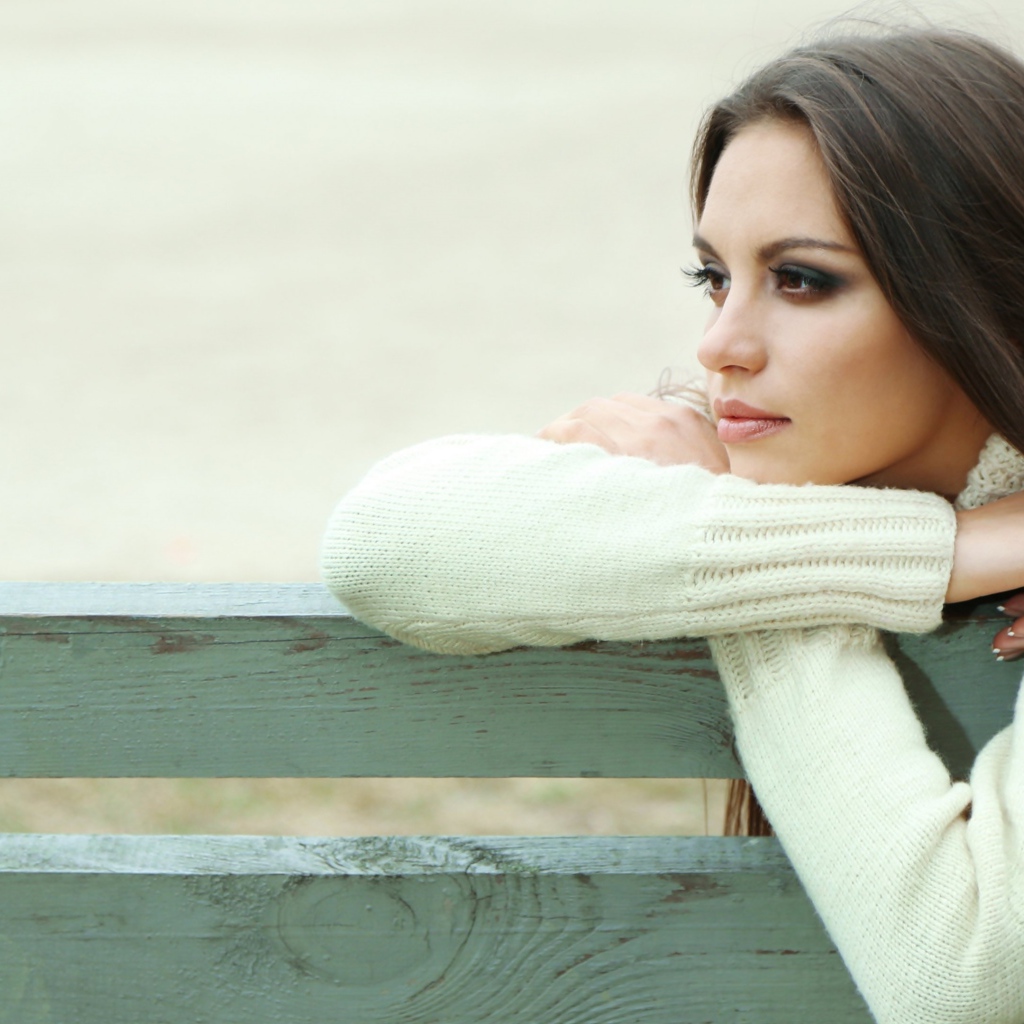 Задумчивая девушка брюнетка в белом свитере сидит на лавке