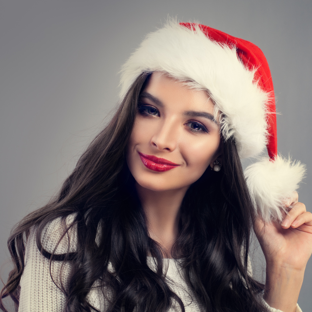 Красивая длинноволосая девушка брюнетка в шапке деда Мороза на сером фоне