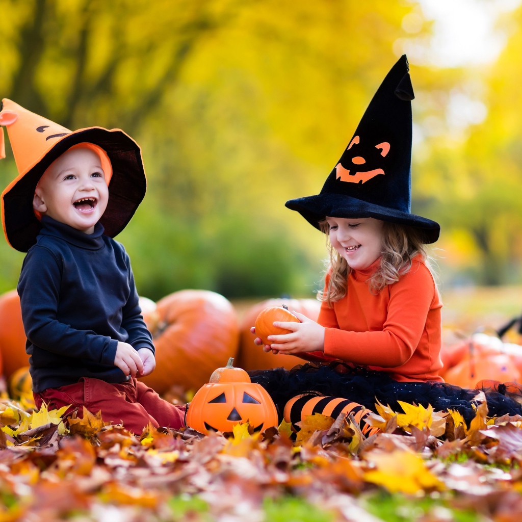Маленькие мальчик и девочка сидят на опавших листьях с тыквами на Хэллоуин
