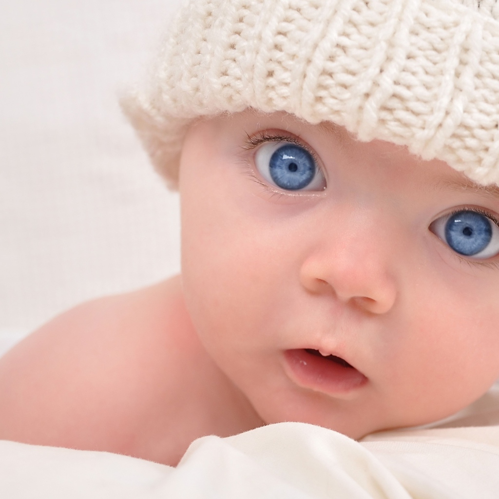 Голубоглазый малыш в белой вязаной шапке на сером фоне