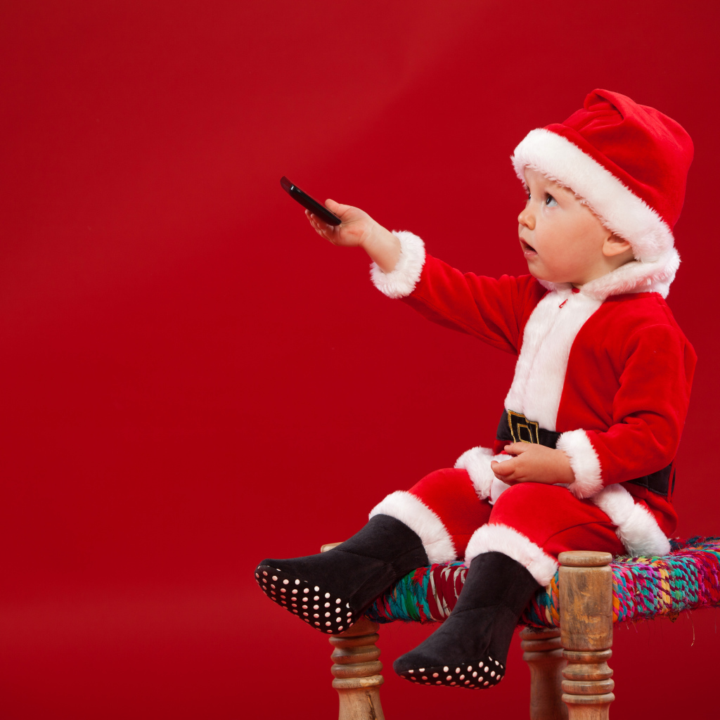 Маленький мальчик в костюме Санта Клауса сидит на стуле на красном фоне