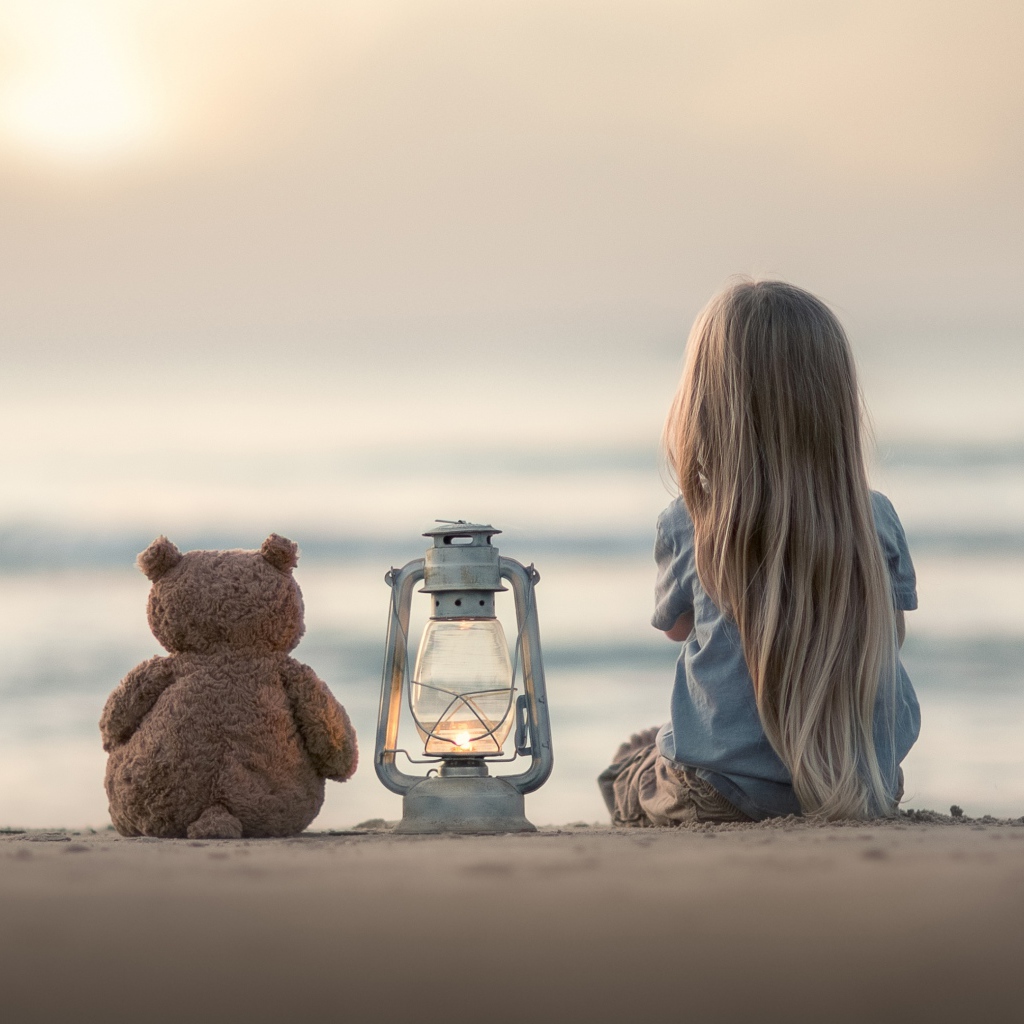 Маленькая девочка сидит на песке с плюшевым мишкой 