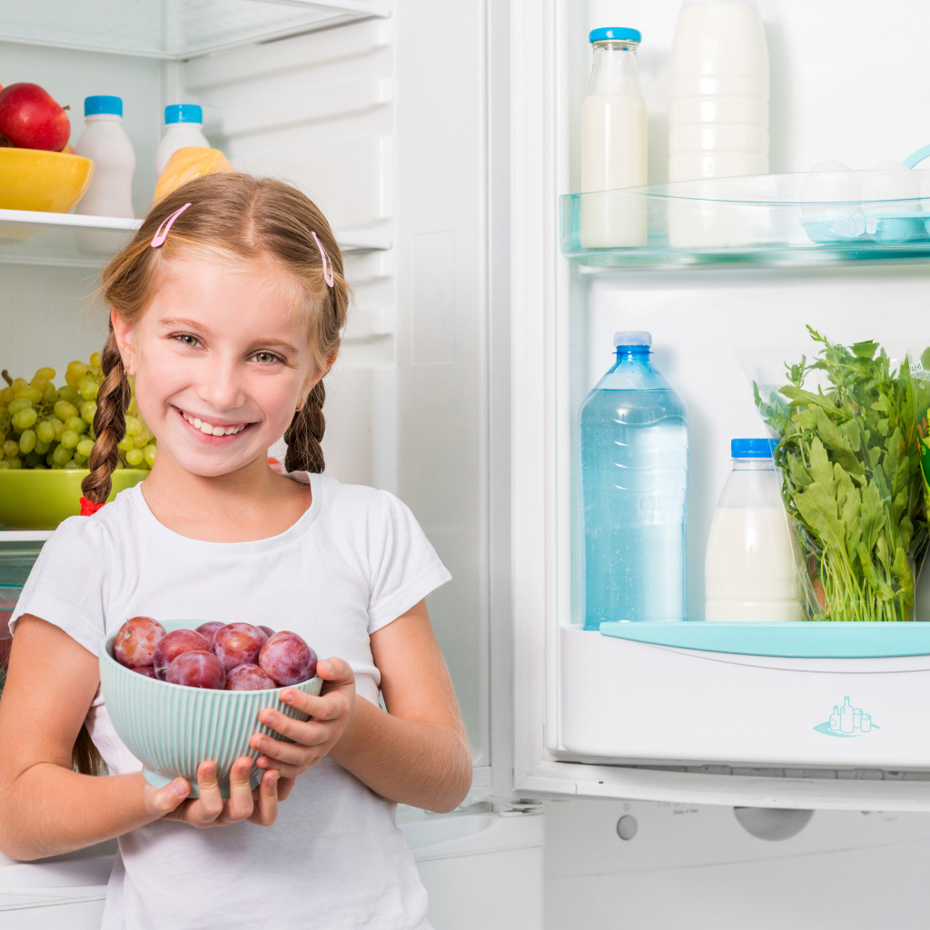 Маленькая девочка с миской со сливами в руках у холодильника 