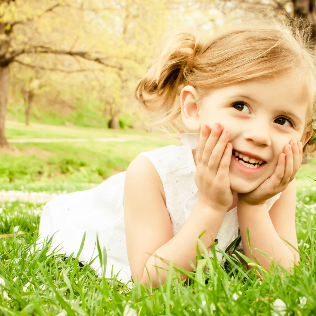 Улыбающаяся девочка в белом платье лежит на зеленой траве