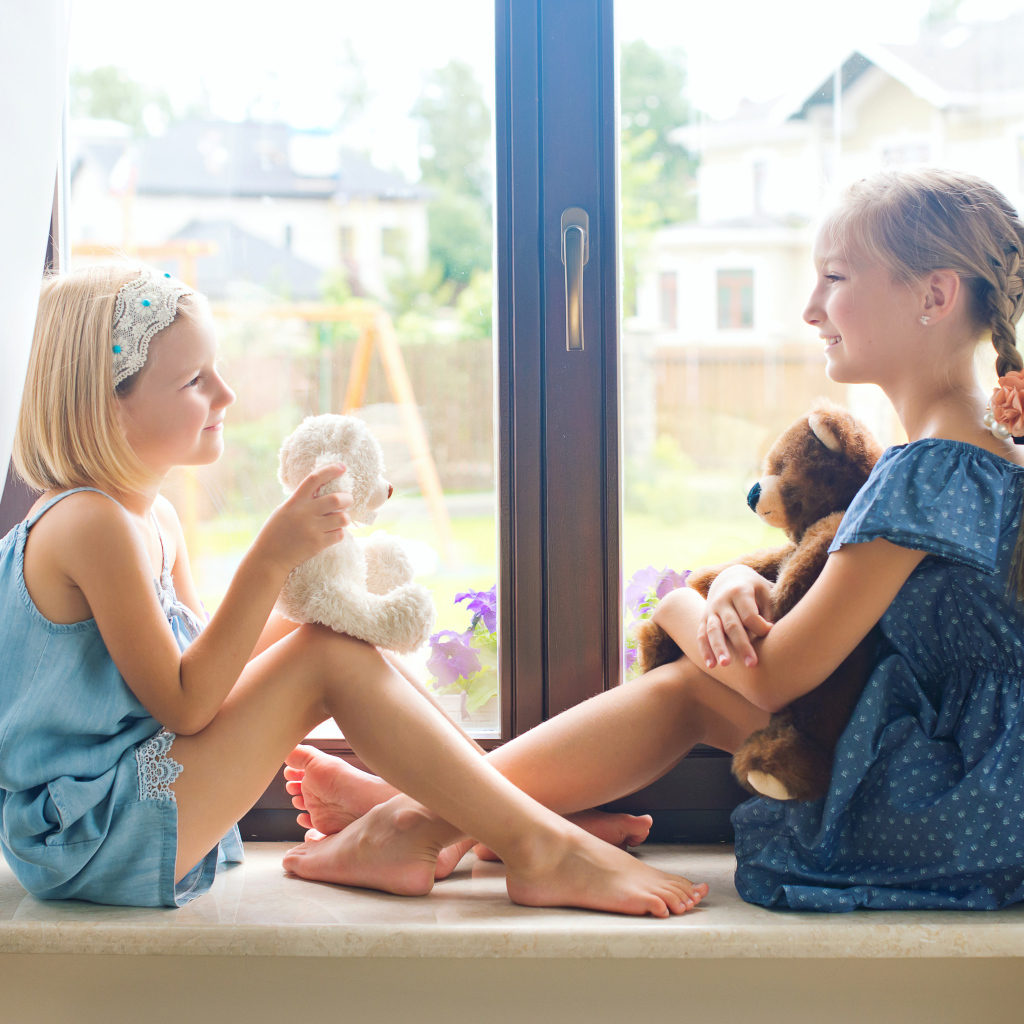 Две маленькие девочки с игрушками сидят на подоконнике у окна