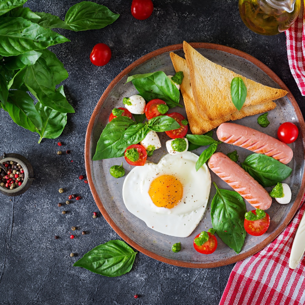 Яичница на тарелке с помидорами, гренками, сыром, сосисками и зеленью базилика