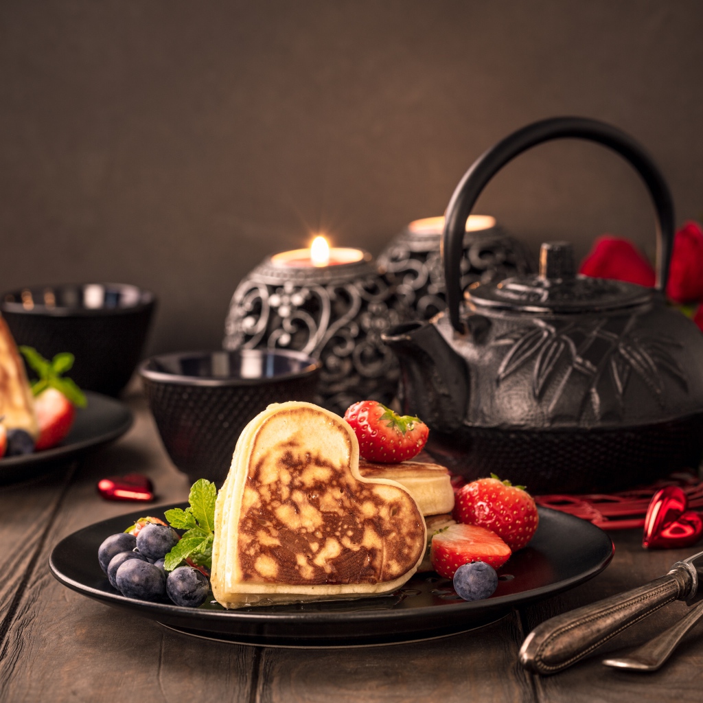 Оладьи в форме сердца с ягодами черники и клубники на столе с чаем и свечами