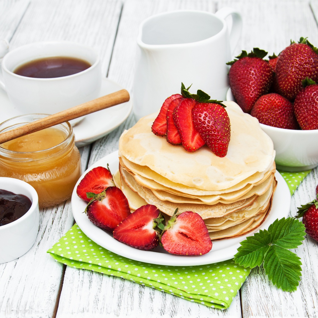 Тонкие блины на столе с ягодами клубники, медом, чаем и джемом