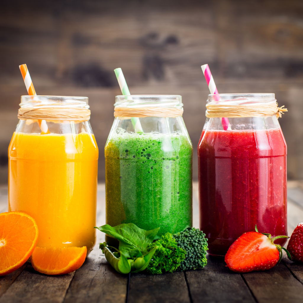 Три банки смузи с трубочкой на столе с апельсином, брокколи и клубникой