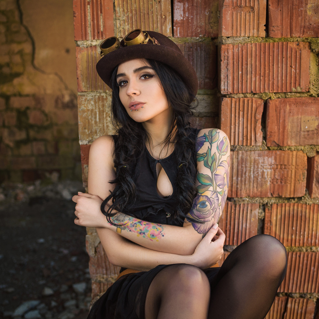The girl in a hat with tattoos on his arm sits near the wall