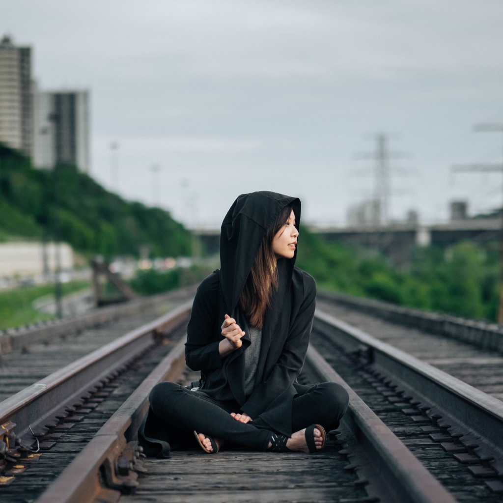 Девушка азиатка в черном балахоне сидит на рельсах 