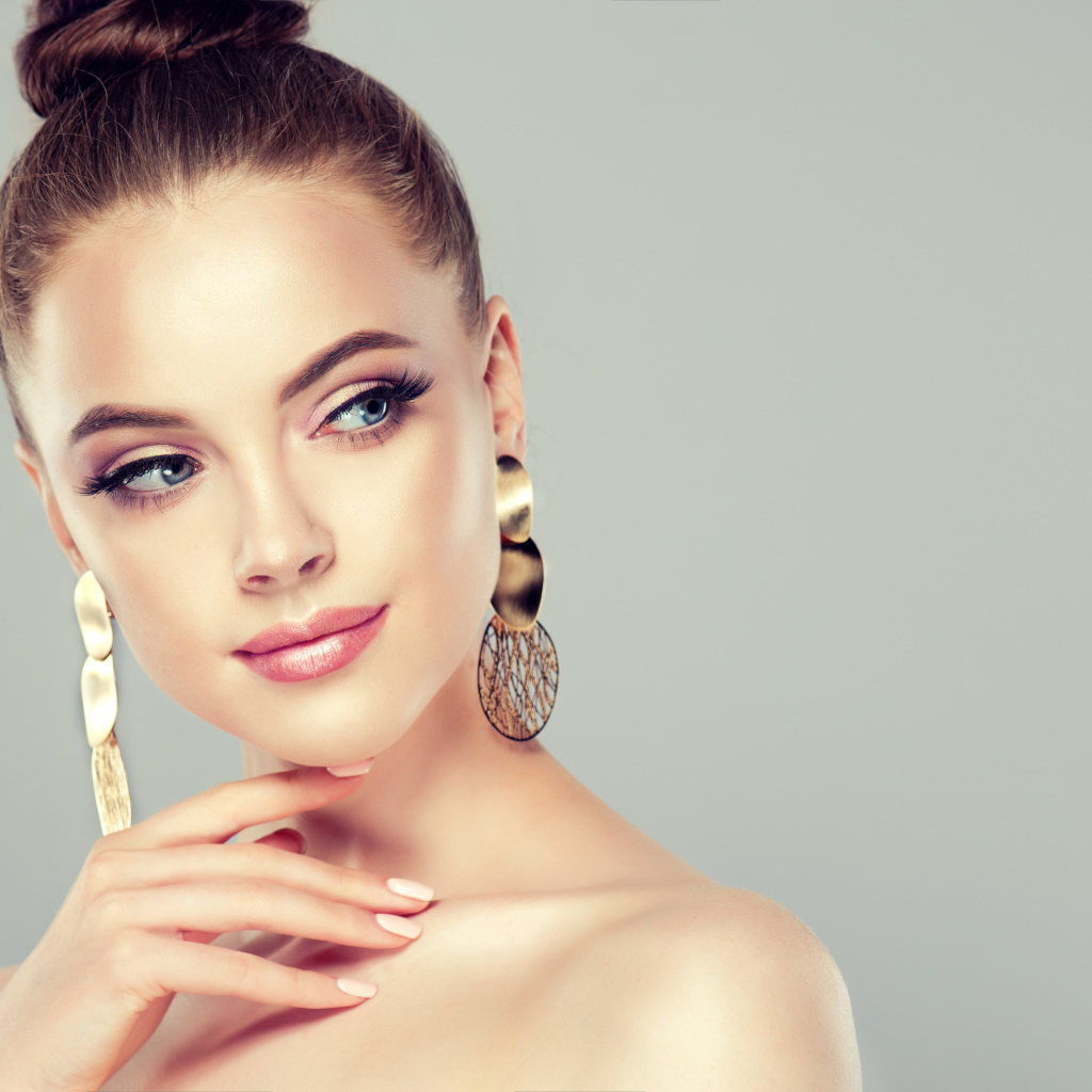 Красивая девушка шатенка с золотыми серьгами в ушах на сером фоне